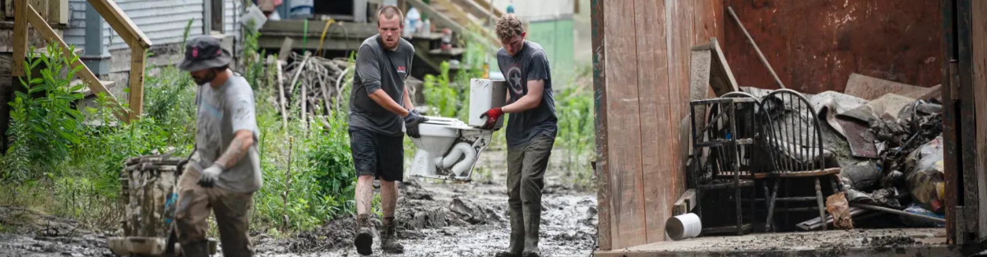 Three men clean up muddy debris outside damaged homes, moving furniture and discarded items into a large dumpster after a flood.